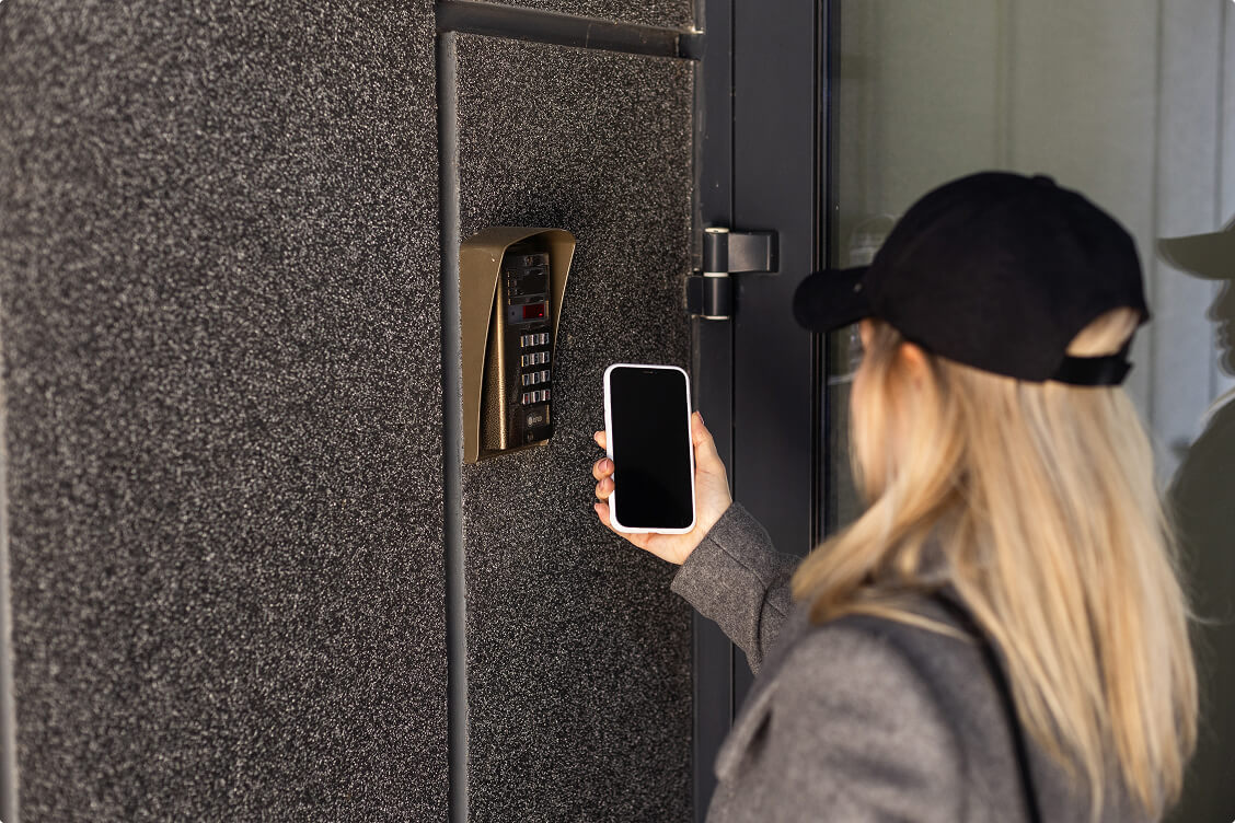 Woman using smartphone for contactless access at a secure building entrance with digital keypad and intercom system.