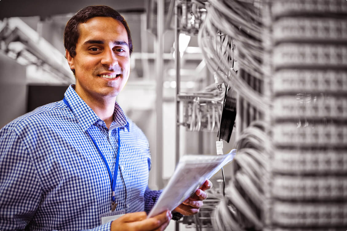 Smiling IT technician holding documents while working in a server room with organized network cables.